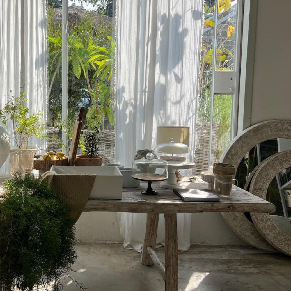 שולחן אוכל מעץ בגימור Whitewash משני גדלים של Fine Lab Home ניצב לצד חלון עם וילונות לבנים שקופים, מלא צמחים, כלי זכוכית, צלחות, כוסות וסטנד לעוגה – כולו מואר באור טבעי.