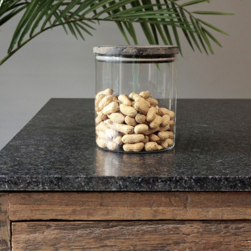 צנצנת זכוכית עם מכסה עץ - בשלושה גדלים מבית Fine Lab Home, מלאה בבוטנים לא קלופים, ניצבת על משטח גרניט כהה ליד מגירת עץ, כשברקע צמח.