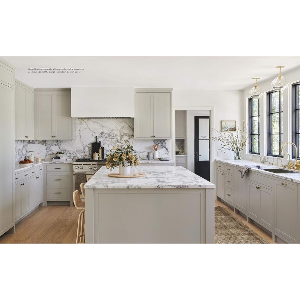 מטבח מודרני עם ארונות אפורים בהירים, משטחי שיש, אביזרי זהב, אי פרחוני וחלונות גדולים המוארים באור השמש — מושלם לחובבי השראה לעיצוב הבית ולאוהבי ספר העיצוב של Fine Lab Home, שנוצר למען החיים.