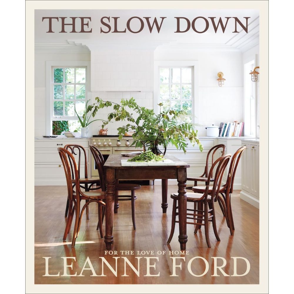 עטיפת הספר "The Slow Down" מאת Fine Lab Home: מציגה שולחן אוכל מעץ עם מרכז שולחן מעוטר בעלים וכסאות במטבח לבן בוהק — השראה לחובבי עיצוב הבית.