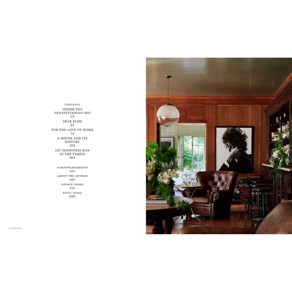 סלון מודרני עם קירות מחופים עץ, כורסה מעור, צמחים ירוקים וספר העיצוב The Slow Down של Fine Lab Home על השולחן; דיוקן מונוכרומי גדול תלוי בסמוך, וטקסט מופיע משמאל לתמונה.