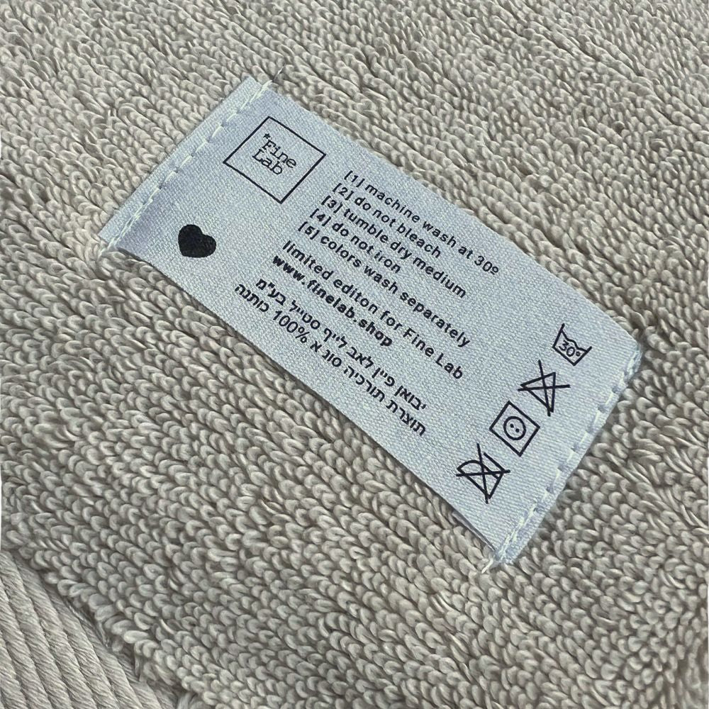 תווית הטיפול בבד על מארז 6 מגבות ידיים - צבע חול של Fine Lab Home, העשויות מכותנה סרוקה, מציגה הוראות כביסה וסמלים לכביסה באנגלית ובעברית, עם סמל לב - אידיאלי למגבות יוקרה או מגבות ידיים.