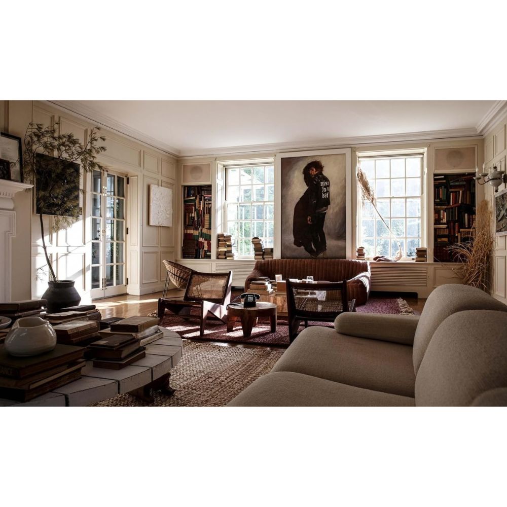 סלון מואר בשמש, בעיצוב ליאן פורד, כולל מדפי ספרים, דיוקן גדול בין החלונות, פינות ישיבה נעימות, שולחנות קפה וערימות ספרים — כולל ספר העיצוב The Slow Down מאת Fine Lab Home — על הרצפה ובשולחנות הצד.