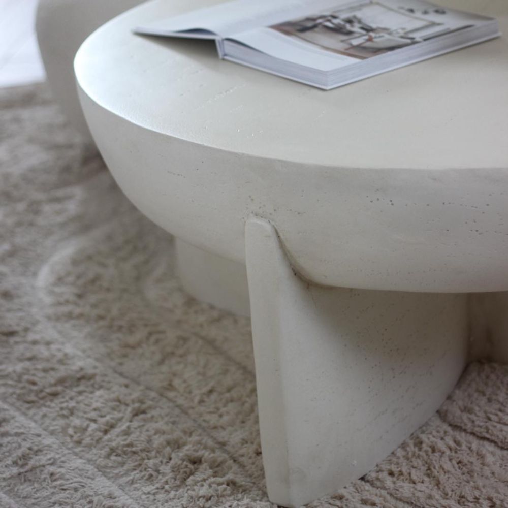 שולחן סלון עגול דמוי אבן של Fine Lab Home עם רגליים עבות, מונח על שטיח בז' מרקם, ועליו מונחת מגזין פתוח.