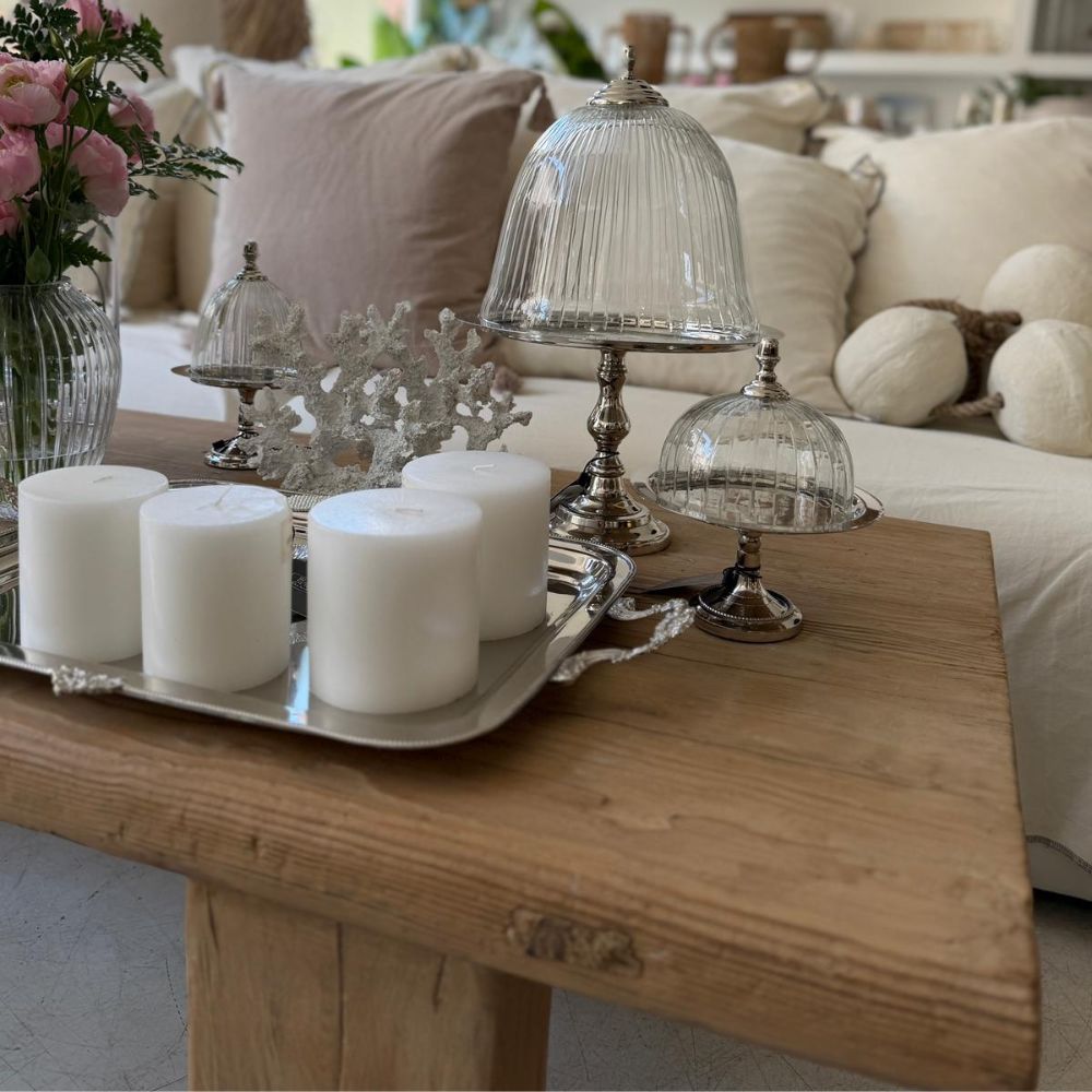 שולחן קפה מעץ, שעליו מונח נר גליל לבן של Fine Lab Home על מגש כסף, כיפות זכוכית עם בסיסים מכסף ופרחים ורודים, יוצר סלון מזמין, המושלם עבור נרות לעיצוב הבית.