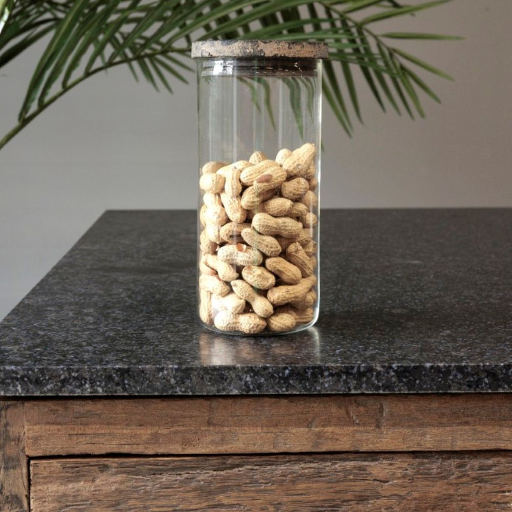 צנצנת זכוכית עם מכסה עץ - בשלושה גדלים מבית Fine Lab Home, מלאה בבוטנים לא קלופים, ניצבת על משטח אבן כהה ליד מגירת עץ, כשברקע נראים עלי צמח ירוקים.