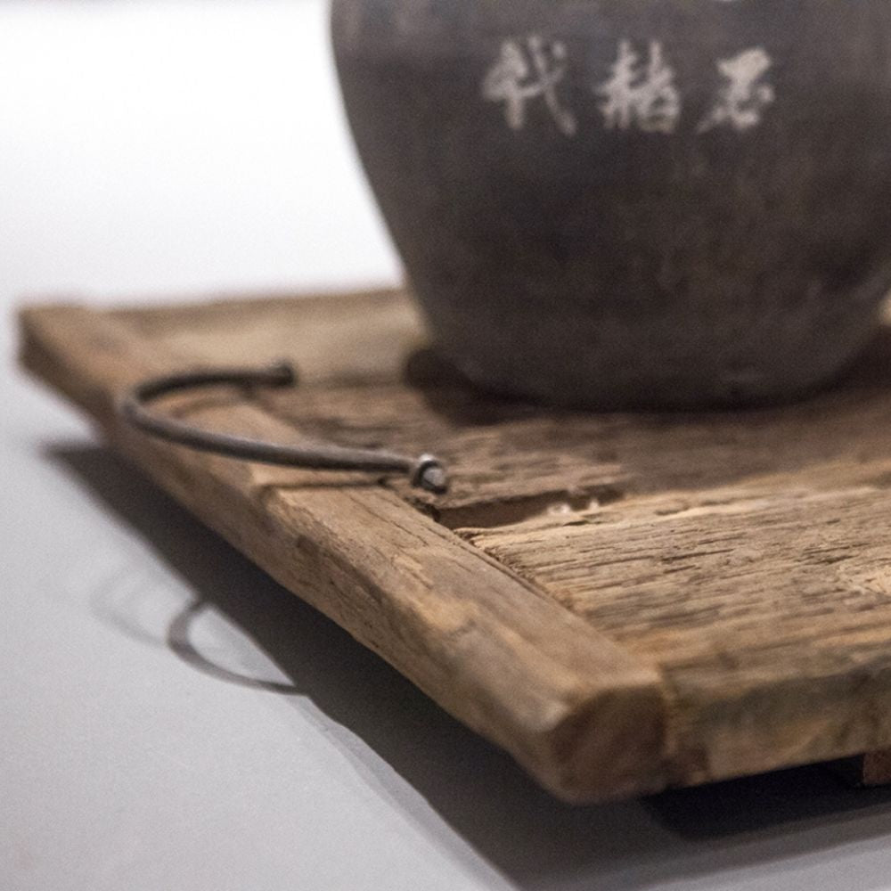 תקריב של מגש עץ עתיק עם ידיות מבית Fine Lab Home, הכולל ידית מתכת ועליו עציץ קרמיקה עם סימניות סיניות לבנות.