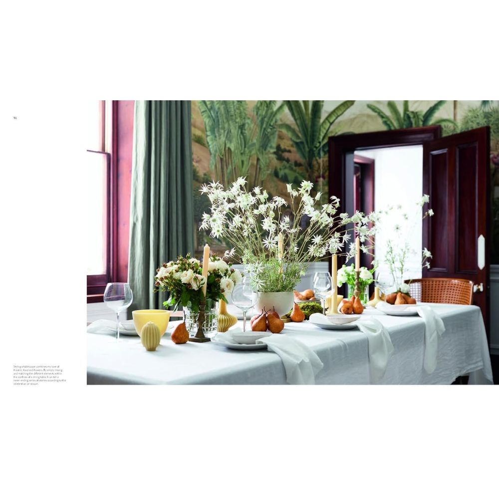 שולחן אוכל מעוצב במפות לבנות ונרות מעורר את רגעי ספר עיצוב של Fine Lab Home בבית, המוצב ליד חלון עם וילונות ירוקים וציור קיר בוטני ברקע.