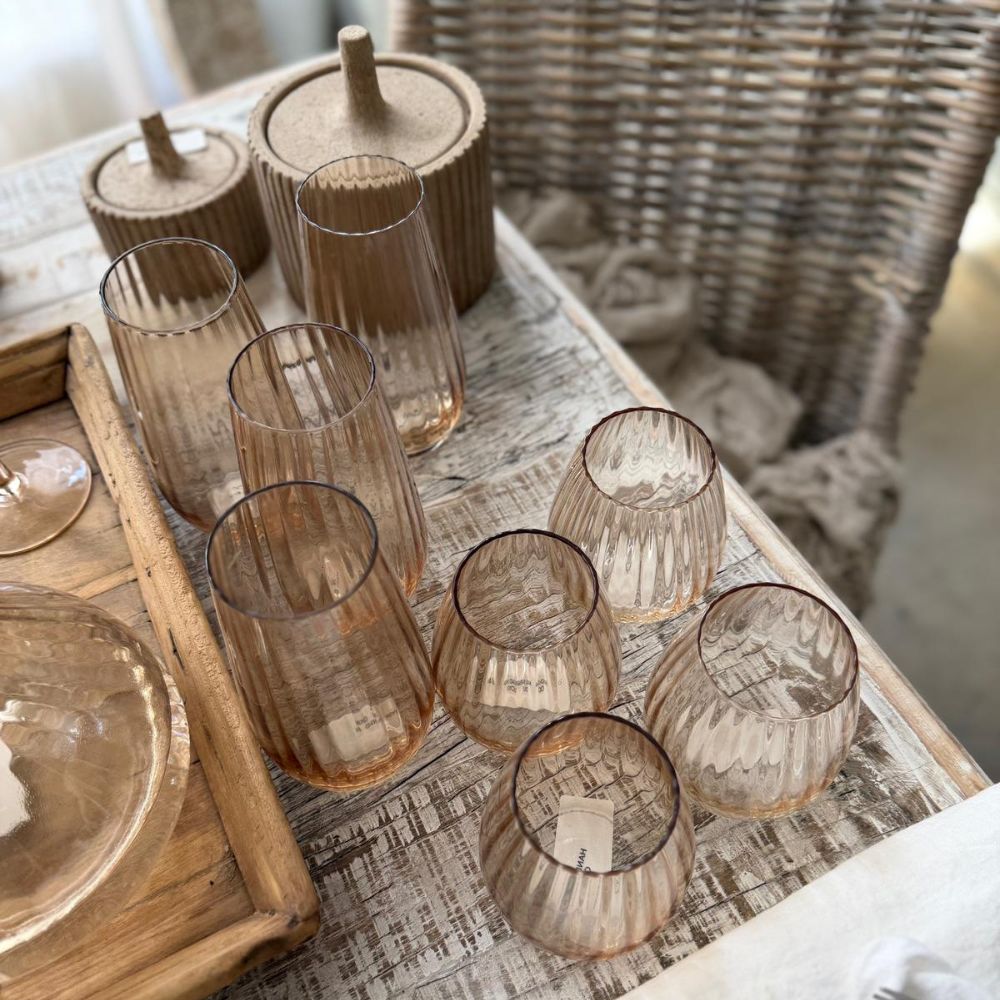 סט של שש כוסות זכוכית גבוהות בגוון שמפניה של Fine Lab Home מסודר על שולחן עץ כפרי ליד שני מיכלים עם מכסה וכסא קלוע.