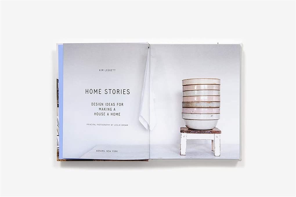 ספר פתוח עם ספר עיצוב סיפורי בית מאת Fine Lab Home בעמוד השמאלי וקערות מוערמות על שרפרף בעמוד הימני, המעוררים השראה לעיצוב הבית וסגנון אישי.