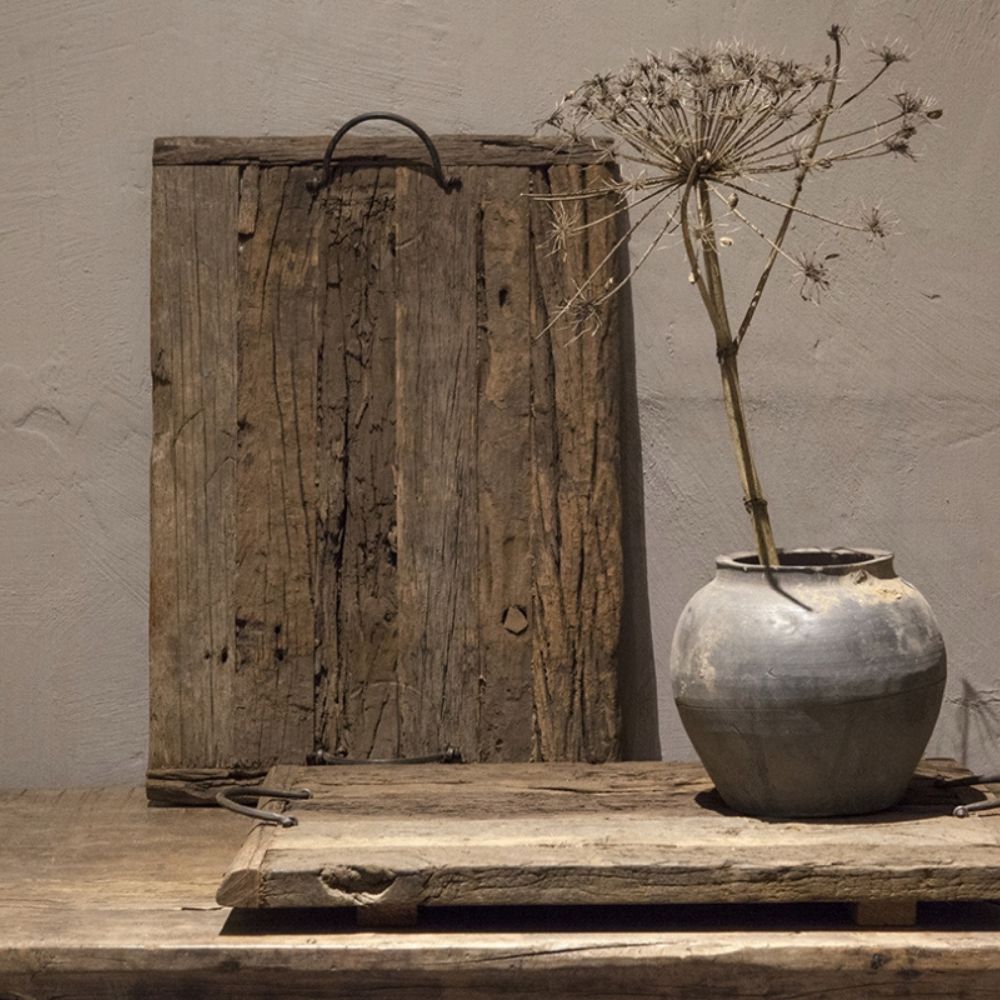 אגרטל קרמיקה אפור עם פרח מיובש ניצב על משטח עץ כפרי לצד מגש עץ עתיק עם ידיות של Fine Lab Home, כשמאחוריו מונחות שתי לוחות עץ בלויות על רקע קיר ניטרלי.