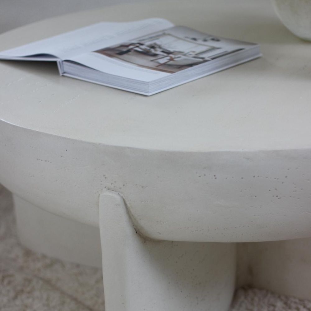 The Fine Lab Home שולחן סלון עגול דמוי אבן הוא שולחן קפה לבן ועגול עם רגליים עבות ועיצוב מינימליסטי, המוצג כאן עם מגזין פתוח על משטחו.