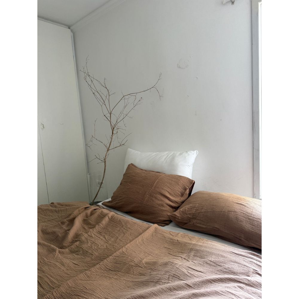 A Fine Lab Home סט מצעים טטרה זוגי חום – סט מצעים למיטה ברוחב 1.60 מטר בצבע חום, בשילוב עם כרית לבנה וענף חשוף בפינה על רקע קיר לבן חלק.