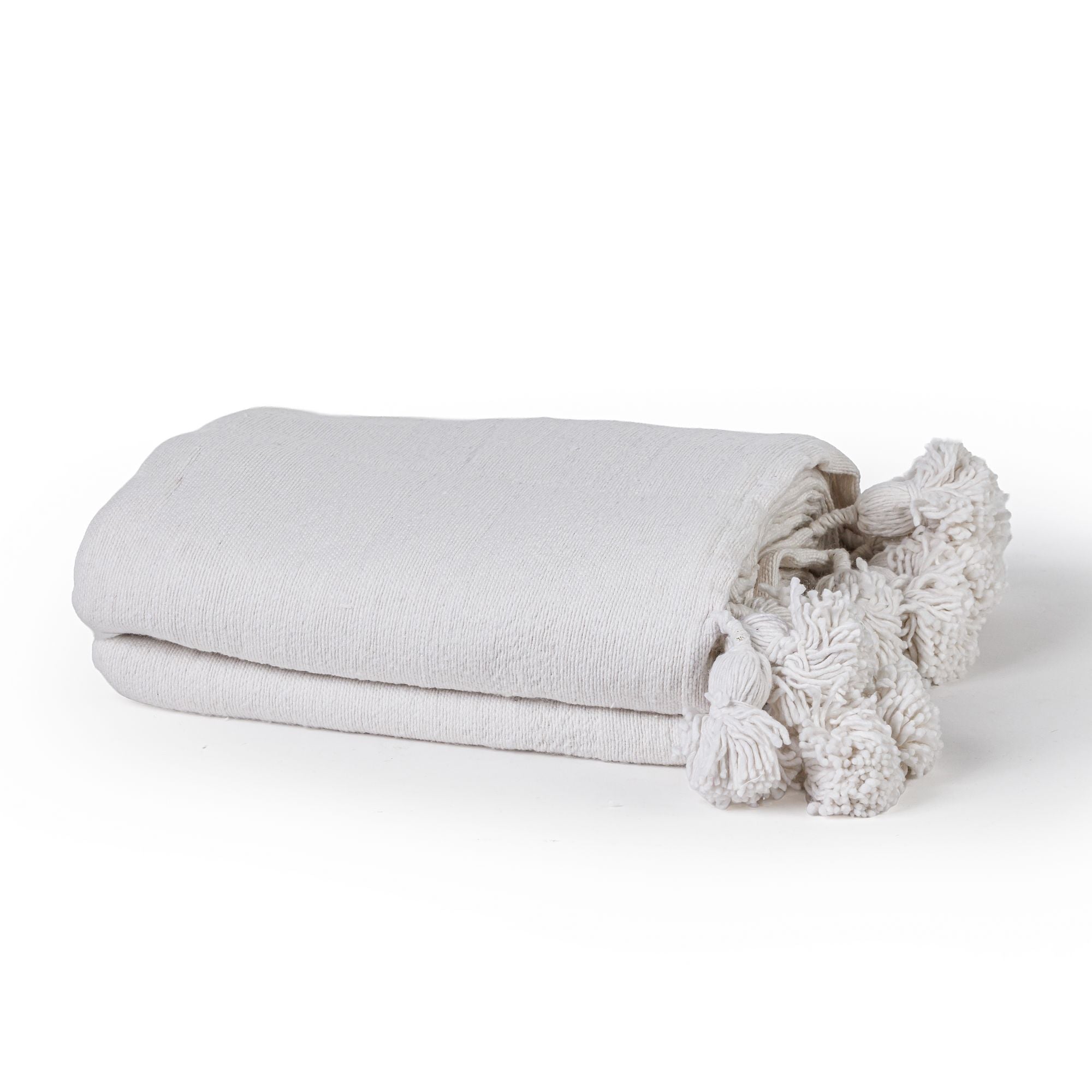 שמיכת מרקש מקופלת עם פונפונים מבית Fine Lab Home, העשויה מבד מרקם בגוון לבן-שבור, עם ציציות דקורטיביות בקצה אחד בסגנון בהשראה מרוקאית, על רקע לבן חלק.