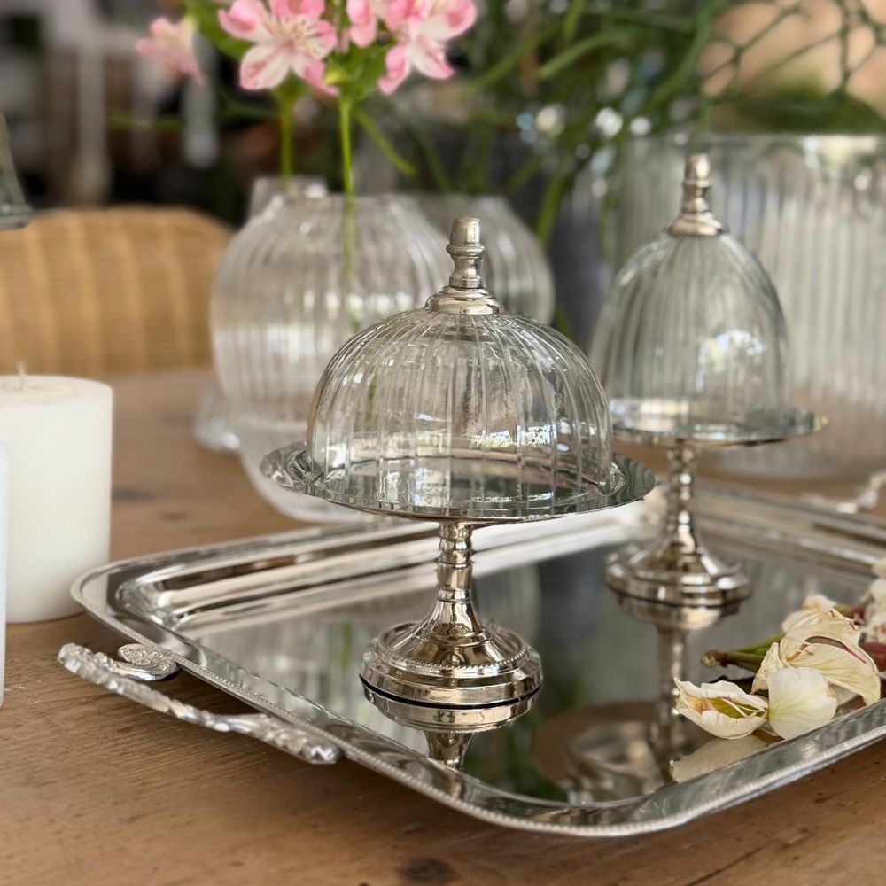 פעמון זכוכית וכסף אלגנטי מבית Fine Lab Home, זמין בשלושה גדלים, מונח על מגש מבריק לצד נר, פרחים ואגרטלי זכוכית על שולחן עץ.