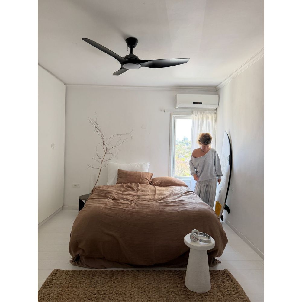 אדם עומד ליד חלון בחדר שינה מינימליסטי עם קירות לבנים, גלשן, מאוורר תקרה, וסט מצעים זוגי טטרה חום של Fine Lab Home – סדין 1.60 ומפה.