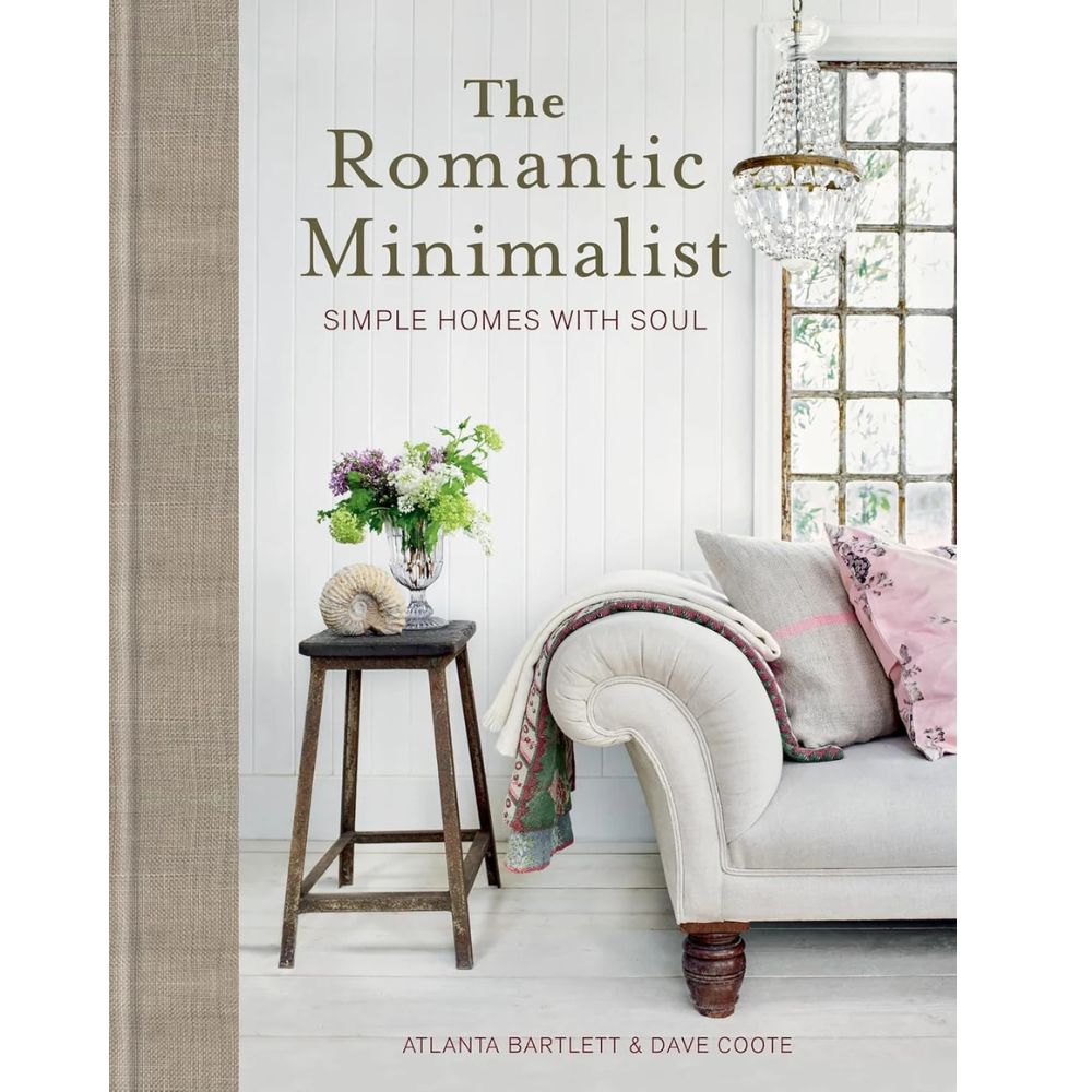 כריכת הספר "The Romantic Minimalist: Simple Homes with Soul" מאת Fine Lab Home, המציגה ספה לבנה, שרפרף עץ עם פרחים וקונכייה, וחלונות בחדר מינימליסטי בהיר בגוונים ניטרליים.
