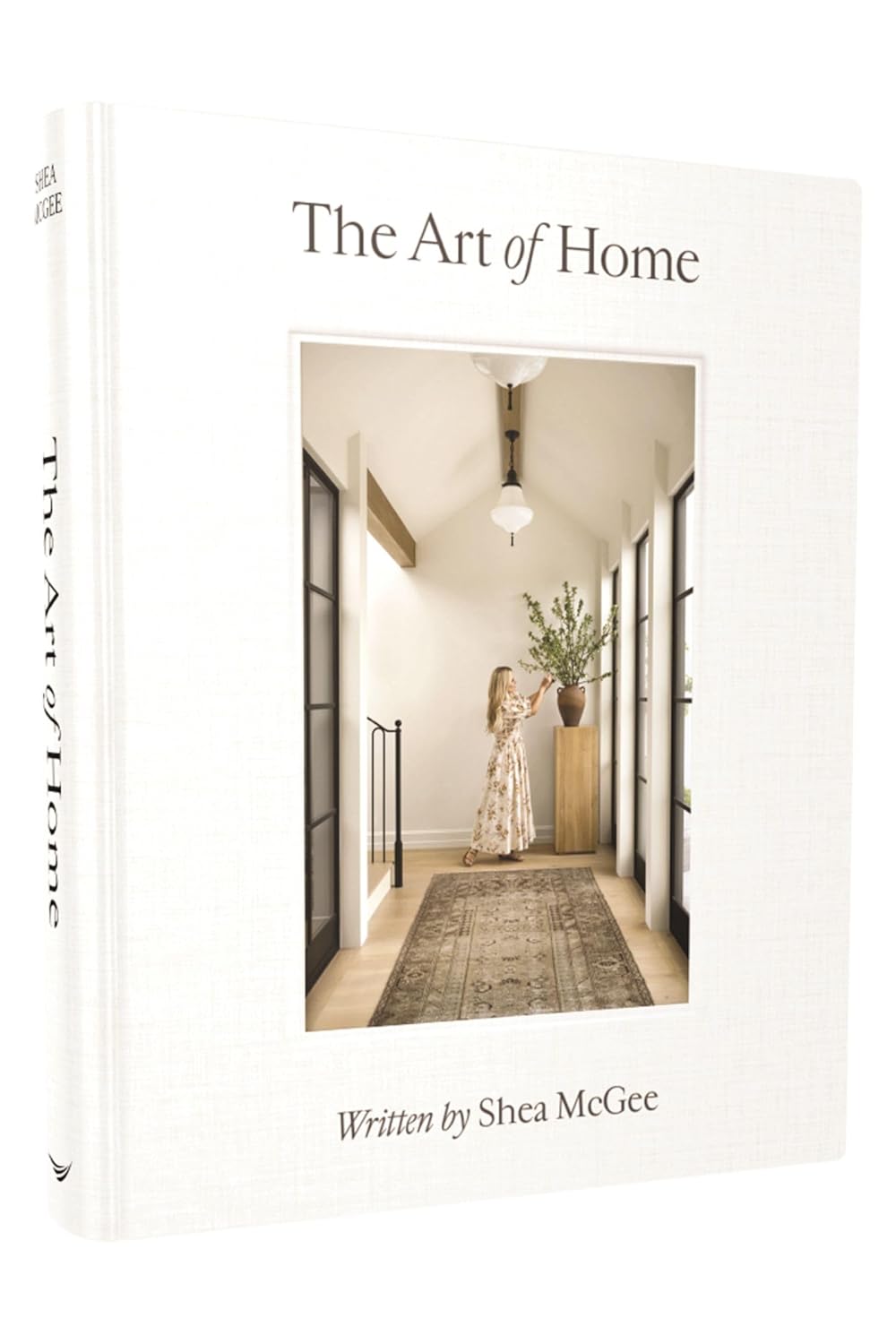 על עטיפת הספר "The Art of Home" מבית Fine Lab Home מופיעה אישה המסדרת פרחים במסדרון מואר בשמש, המשקפת את סגנון העיצוב האופייני למותג.
