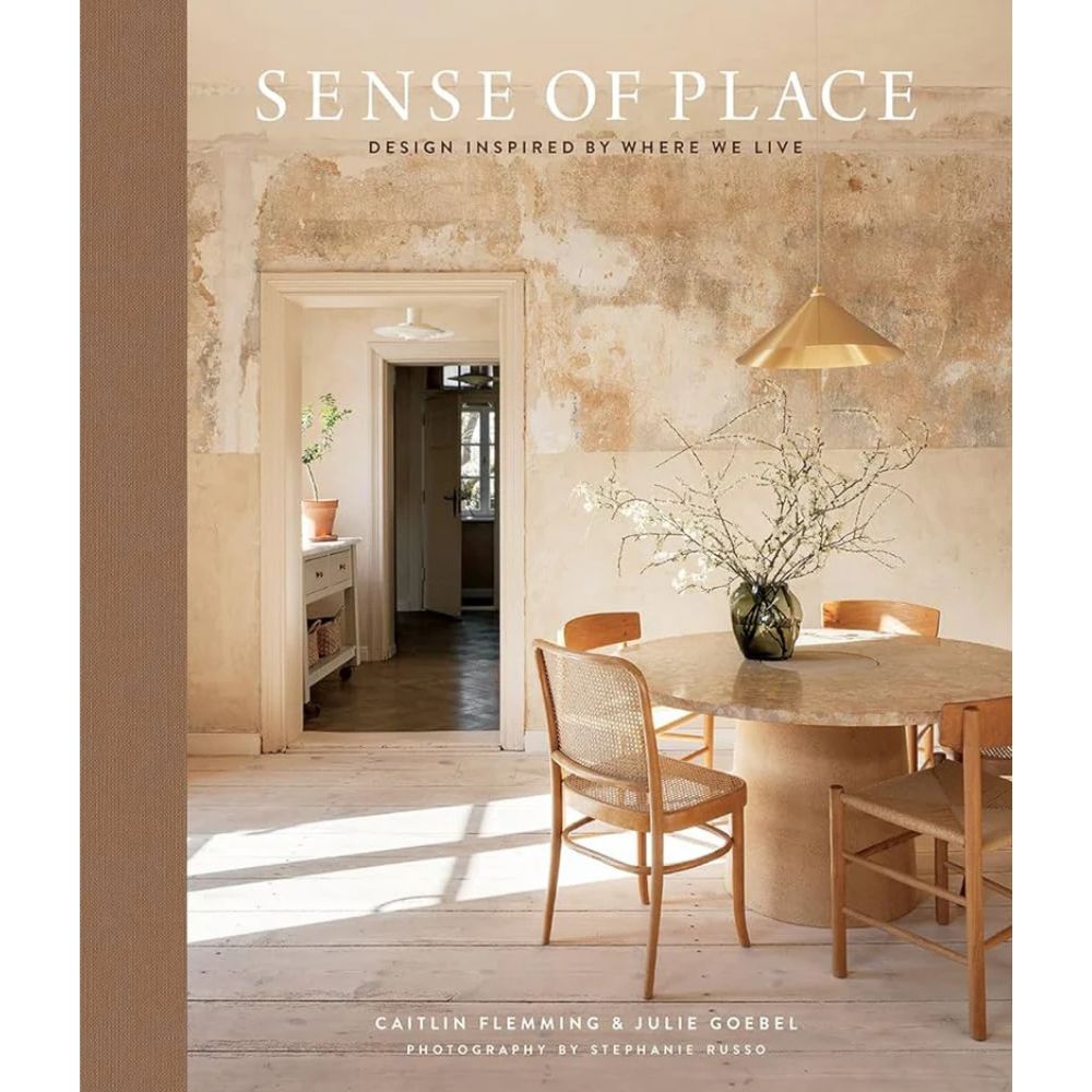 כריכת הספר "ספר עיצוב sense of place" מאת Fine Lab Home מציגה פנים כפרי – שולחן עגול, ארבע כיסאות, אגרטל עם ענפים וקיר בלוי. אידיאלי לאוהבי השראה לבית. הכותרת והמחברים מופיעים בחלק העליון.