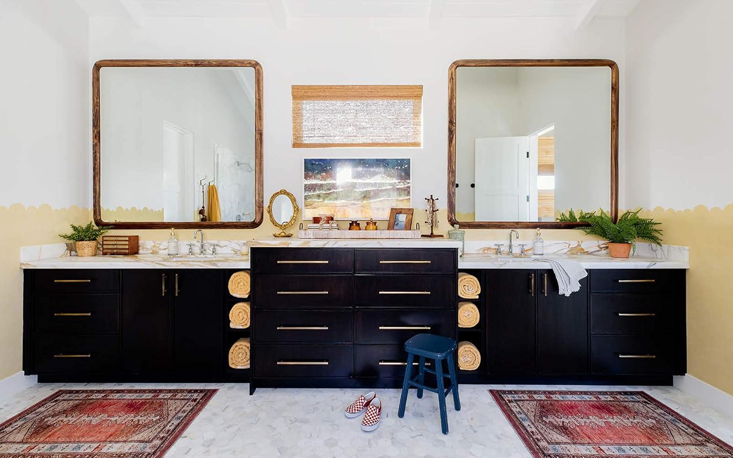 חדר רחצה מודרני עם כיורים כפולים, ארונות כהים, מראות, מגבות מגולגלות, שידה מרכזית, שטיחים אדומים, שרפרף כחול וצמחים – תמונה שנלקחה היישר מתוך סיפור הבית בספר העיצוב של Fine Lab Home.