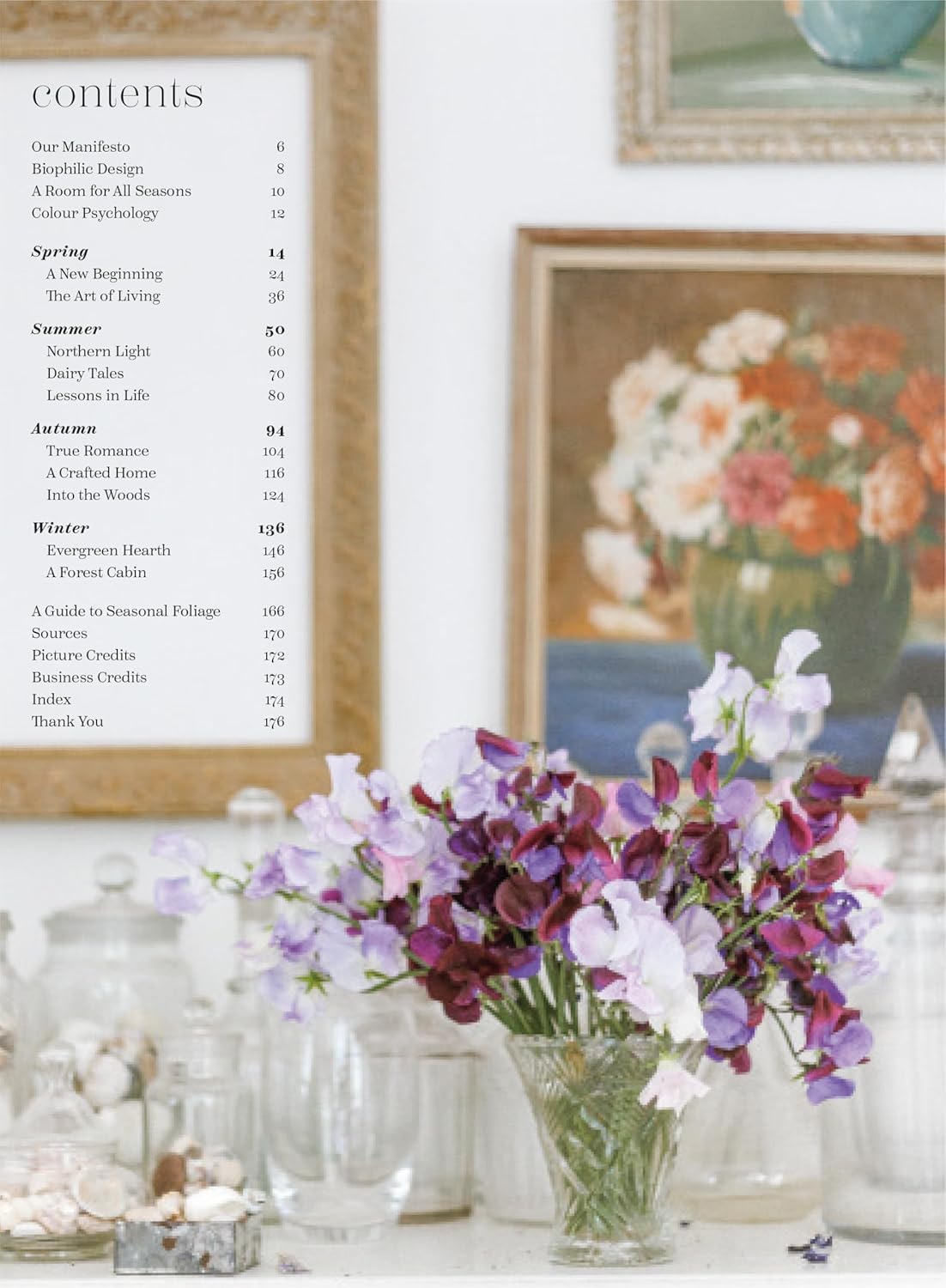 ענף עלים גדול באגרטל קרמי מונח על שרפרף עץ ליד מיטה מסודרת עם מצעים ניטרליים, המקשר בין הטבע לעיצוב הבית; מוצג: ספר עיצוב בית לכל עונות השנה מאת Fine Lab Home.