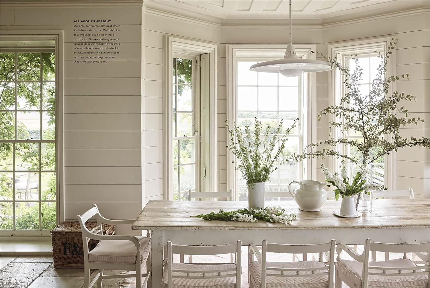 ספר העיצוב the art of living with white מבית Fine Lab Home מציג פינת אוכל מוארת עם שולחן עץ, כיסאות לבנים, סידורי פרחים וחלונות גדולים לנוף ירוק, ליצירת מינימליזם נייטרלי והרבה אור טבעי.