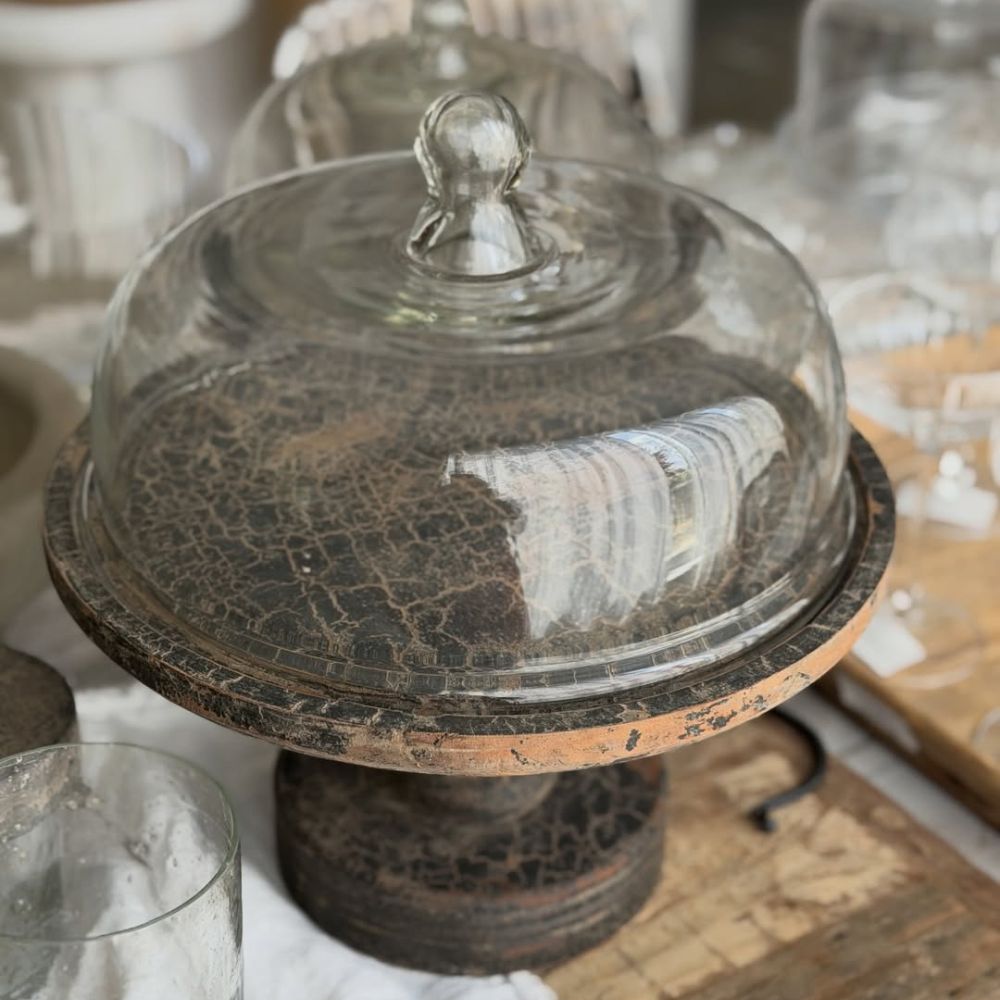 The Fine Lab Home פעמון עוגה על רגל מכסה זכוכית ניצב על גבי בסיס עץ כפרי, וכיפת הזכוכית שלו מכסה מעמד עוגות ריק ומזדקן, המוקף בכלי זכוכית נוצצים אחרים.