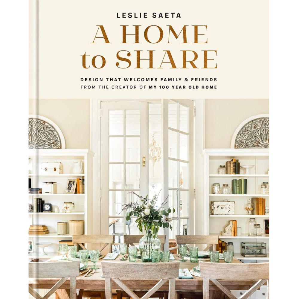 עטיפת הספר "A Home to Share" מאת לסלי סאטה, בהוצאת Fine Lab Home, מציגה שולחן אוכל ערוך עם כלי זכוכית, כסאות עץ ומדפים מובנים בחלל מואר ואלגנטי — השראה מושלמת לכל מי שמתעניין באירוח או בעיצוב פנים.