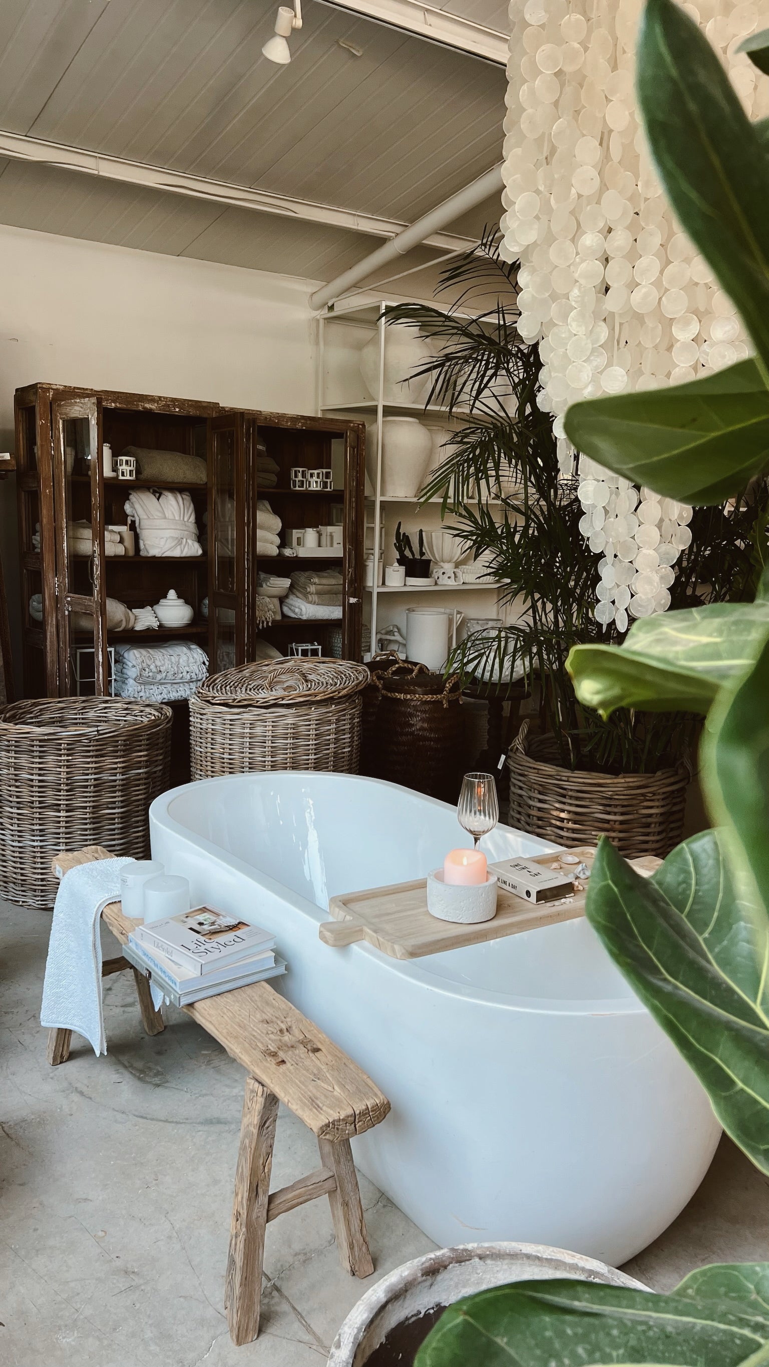 אמבטיה עצמאית עם ספסל עץ ממוחזר ומגש של Fine Lab Home ניצבת בחדר נעים ומלא בסלי נצרים, מדפי מגבות, צמחים ונברשת פגזים תלויה.