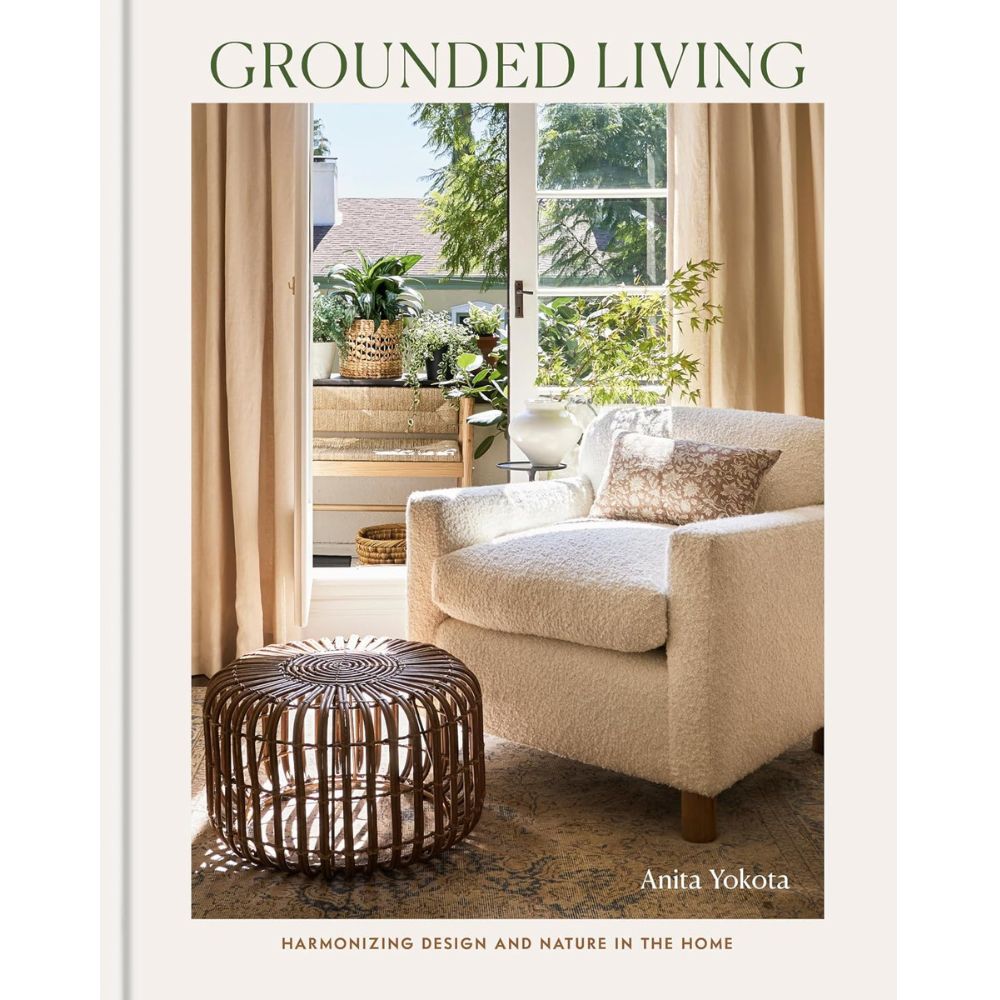 עטיפת הספר “Grounded Living” מבית Fine Lab Home, המציגה כורסה נעימה, הדום עגול מקש וצמחייה שופעת בחדר מואר באור השמש, עם וילונות בצבע בז' ונוף של עצים בחוץ — חלל מזמין הממחיש להפליא את העיצוב הביופילי ואת השימוש בחומרים טבעיים.