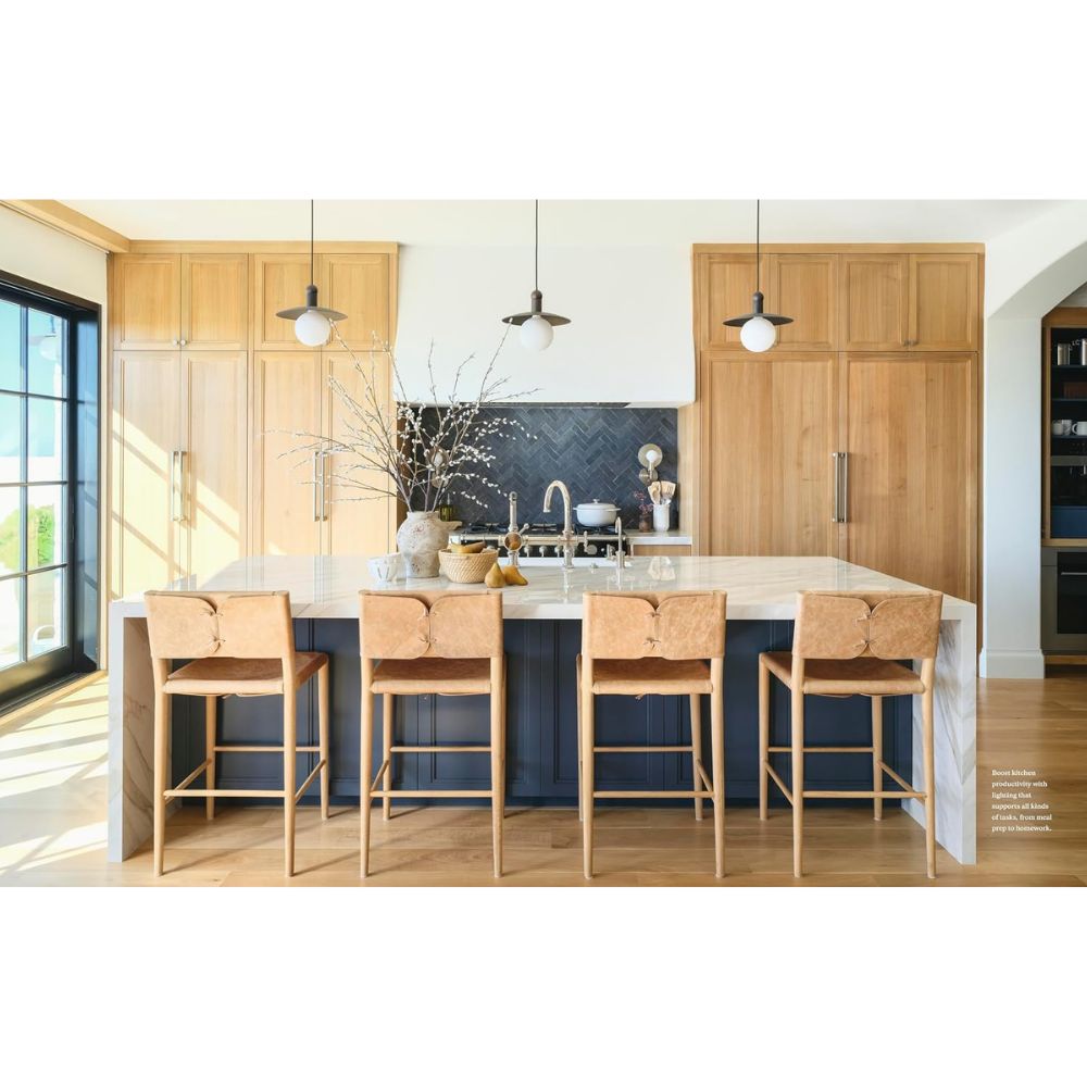 מטבח מודרני עם ארונות מעץ בהיר, אי משיש לבן, ארבעה כסאות בר בצבע חום בהיר, בסיס בצבע כחול כהה, מנורות תלייה ואגרטל עם ענפים על השיש — בהשראת ספר העיצוב “Grounded Living” מבית Fine Lab Home, המדגיש את עיצוב הפנים ואת השימוש בחומרים טבעיים ליצירת אווירה חמימה ומזמינה.