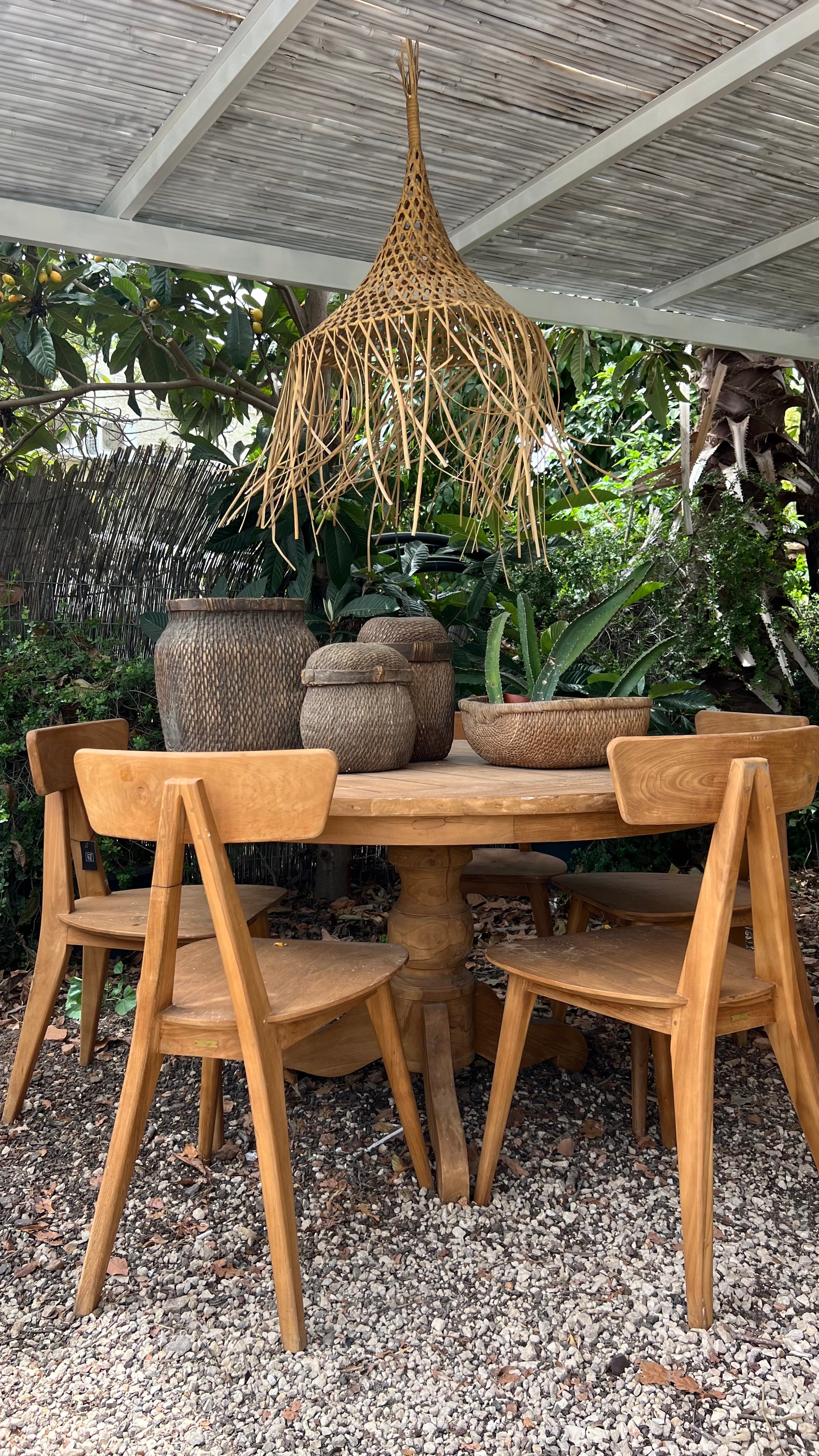 שולחן אוכל עגול-עץ טיק של Fine Lab Home עם ארבעה כיסאות, מעוטר בסלי קש ואגרטלים, ממוקם תחת נברשת ויקטוריאנית בגינה פורחת.