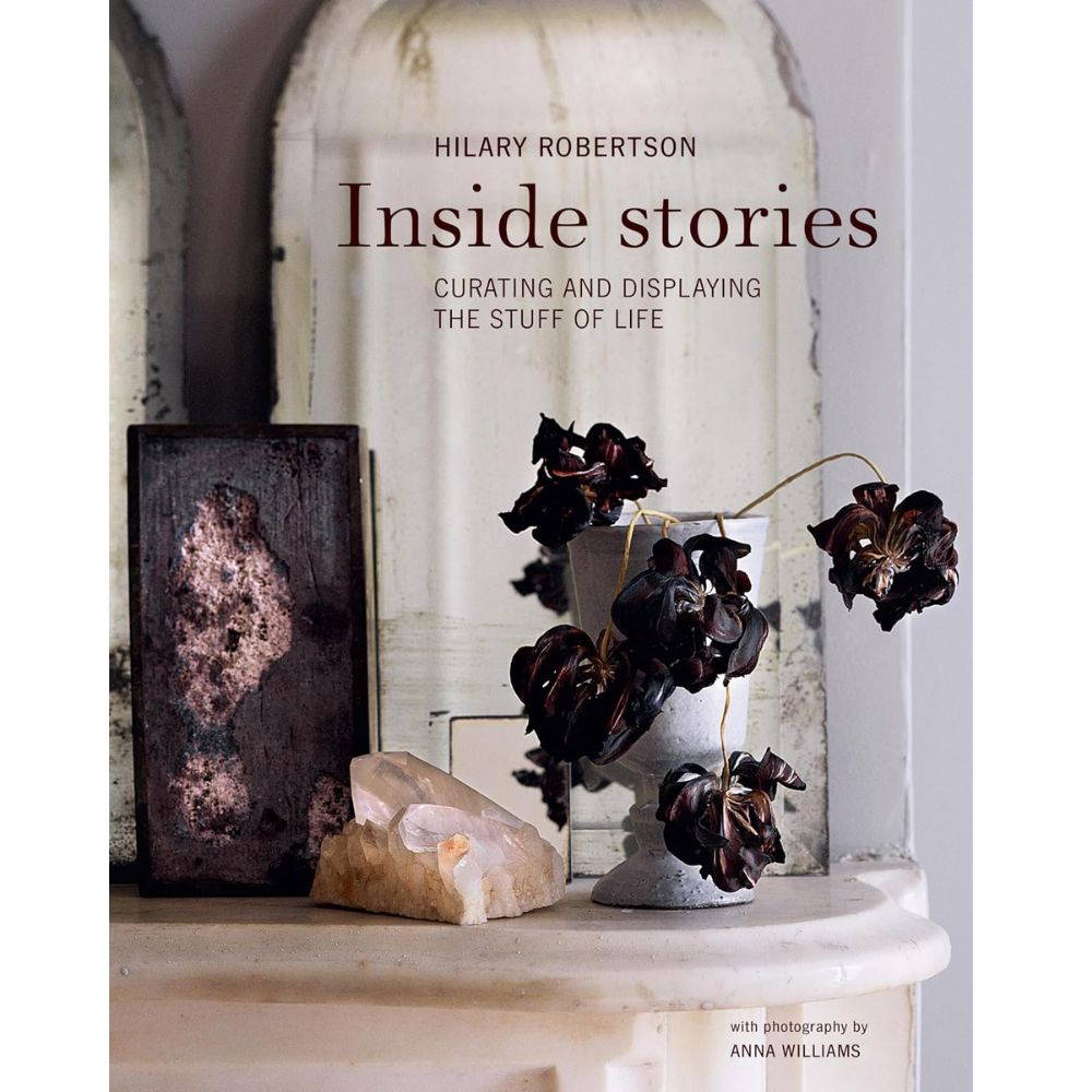 עטיפת הספר "Inside Stories" מאת הילרי רוברטסון בהוצאת Fine Lab Home, המציגה פרחים כהים באגרטל, קריסטל, אובייקט מופשט ואלמנטים דקורטיביים בגוונים ניטרליים — ביטוי מעורר השראה של אוצרות חפצים ועיצוב פנים.