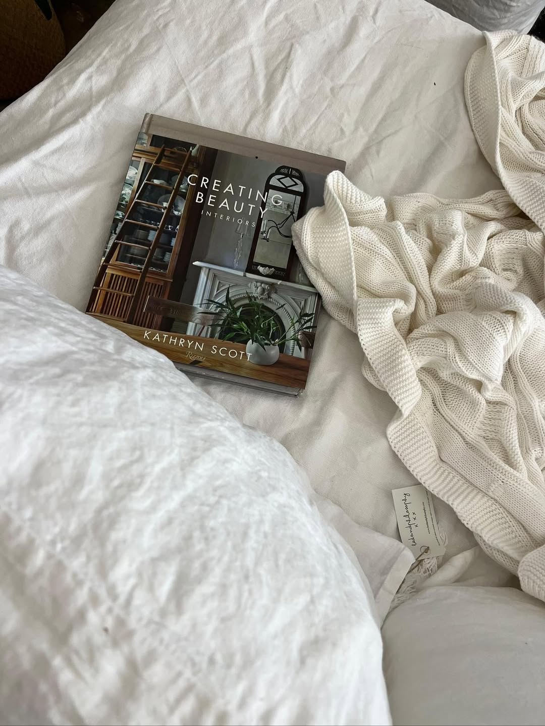 ספר העיצוב "Creating Beauty" מאת Fine Lab Home מונח על כיסוי מיטה לבן לצד שמיכה סרוגה בצבע שמנת וכרית.