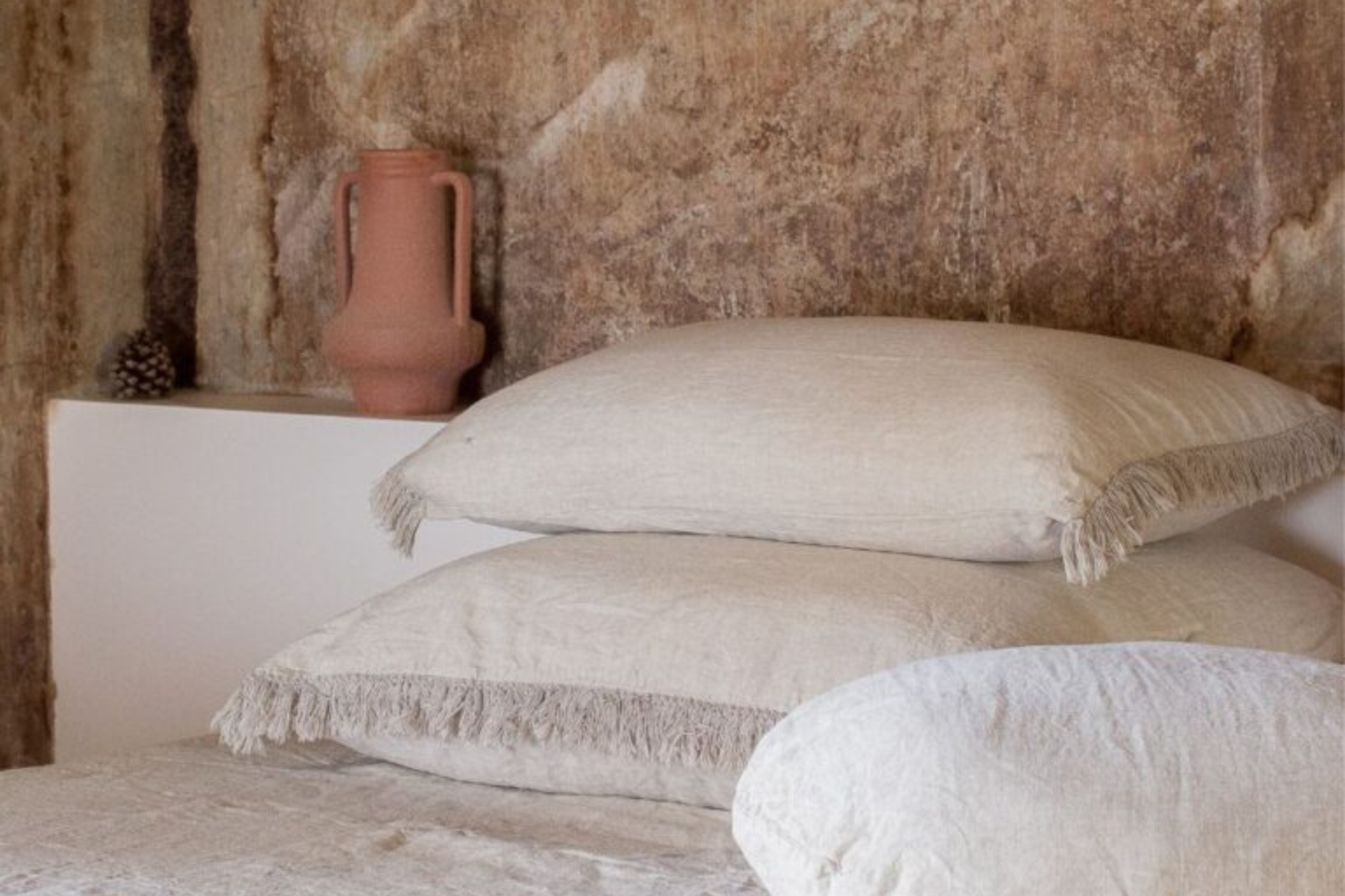 שלוש כריות בצבע בז' של Fine Lab Home כרית פשתן פרנזים מונחות זו על גבי זו על המיטה; אגרטל קרמיקה ורוד ואצטרובל מונחים על מדף לבן על רקע קיר חום מרקם.