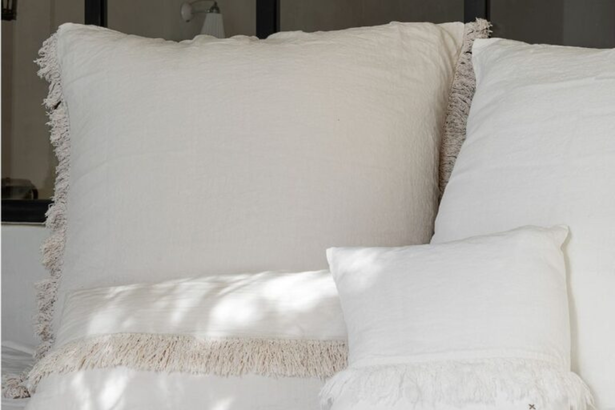 שלוש כרית פשתן פרנזים של Fine Lab Home בצבע לבן, ממגוון גדלים וצבעים, מסודרות על משטח לבן עם אור טבעי המטיל צללים רכים על הכריות היוקרתיות הללו.