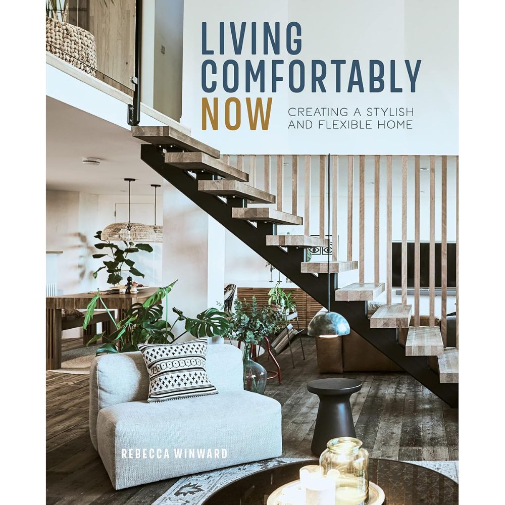 עטיפת הספר "Living Comfortably Now" מבית Fine Lab Home, מאת רבקה ווינוורד, מציגה סלון מודרני עם כורסה אפורה, מדרגות עץ, צמחי נוי ודקורציה אופנתית בסגנון עיצוב הבית. הספר מדגיש פתרונות אחסון חכמים ליצירת חלל אלגנטי ופונקציונלי.