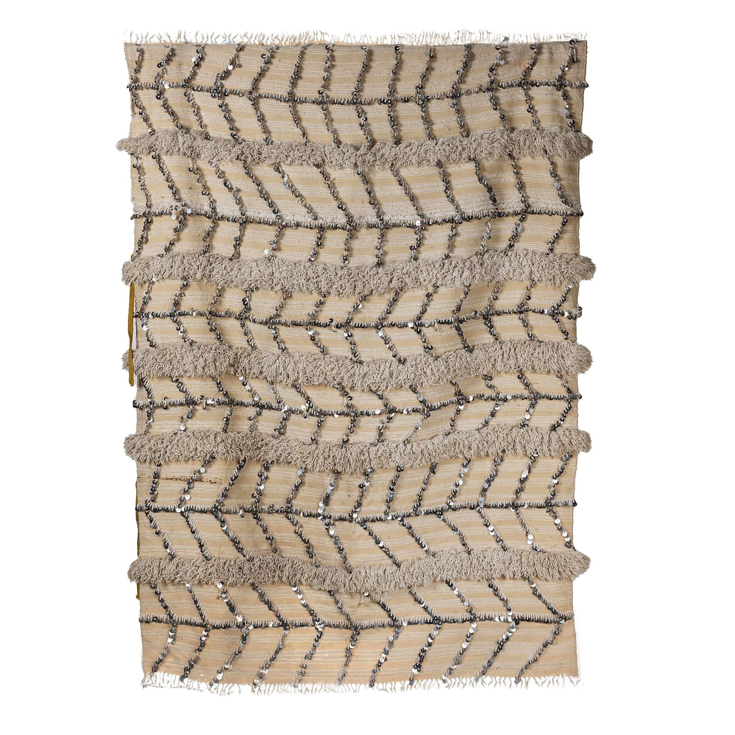 תקריב של שטיח פאייטים מבית Fine Lab Home, עם פסים אופקיים בצבעי בז' וחום בהיר, קווים משולשים ופייטים כסופים בדוגמת שברון, ליצירת מראה אתני אותנטי.