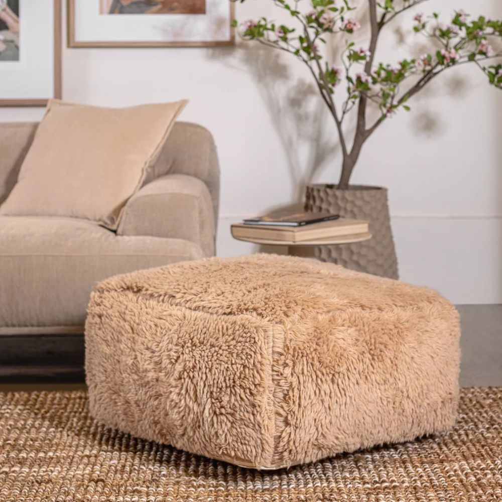 פוף רך בצבע לבן/חול מבית Fine Lab Home מונח על שטיח ארוג מול ספה בצבע בז', ולצידו שולחן צד קטן וצמח פורח בעציץ.
