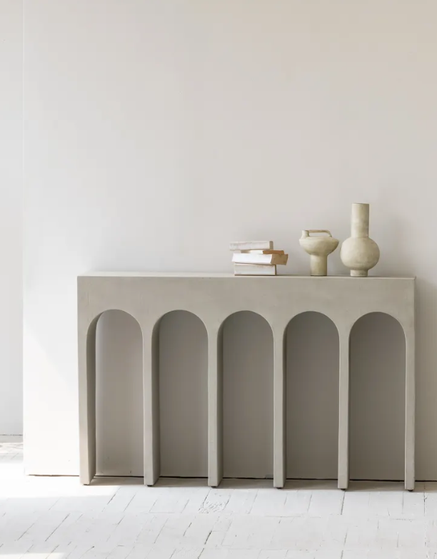 הקונסולה קשתות נורדית מבית Fine Lab Home כוללת חמש רגליים בצורת קשתות ומעוצבת עם ספרים מוערמים ושלושה אגרטלי קרמיקה ניטרליים, הניצבים על רקע קיר לבן חלק.
