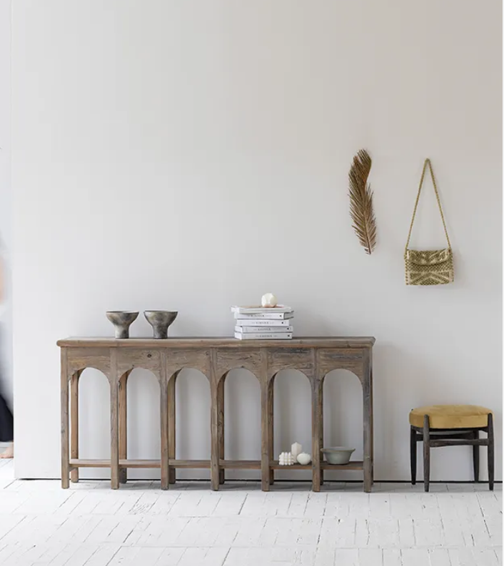 קונסולה נורדית עץ מבית Fine Lab Home ניצבת על רקע קיר לבן, מעוצבת עם שתי קערות, ספרים ערוכים ועיצוב דקורטיבי; לצדהן כיסא בר, תיק תלוי ועלה מיובש המוסיפים נגיעה מודרנית.