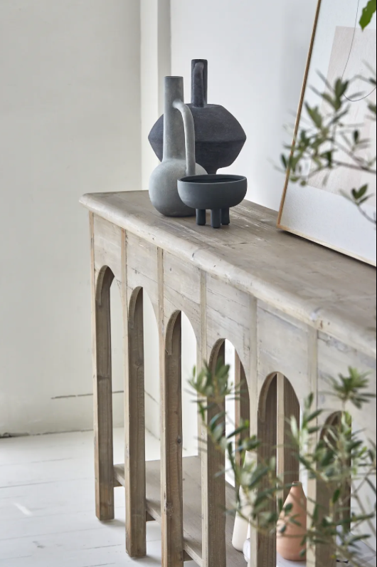 קונסולה נורדית עץ מבית Fine Lab Home ניצבת על רקע קיר לבן, מעוצבת עם שתי קערות, ספרים ערוכים ועיצוב דקורטיבי; לצדהן כיסא בר, תיק תלוי ועלה מיובש המוסיפים נגיעה מודרנית.