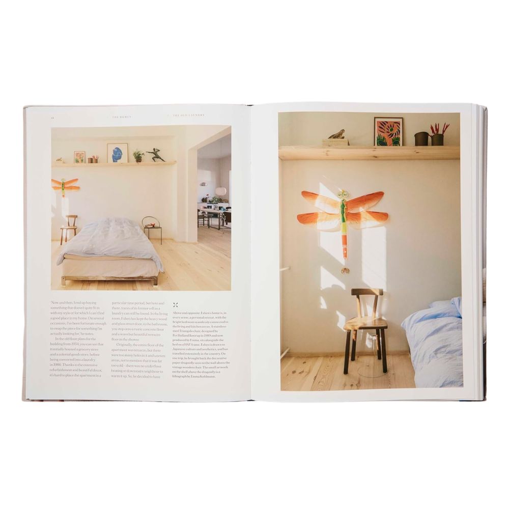 הספר "The Vintage Way" מבית Fine Lab Home מוצג פתוח על תמונה של חדר שינה מינימליסטי עם רצפת עץ בהירה, מיטה פשוטה, כיסא עץ וקישוט כתום — השראה לעיצוב פנים בסגנון סקנדינבי.