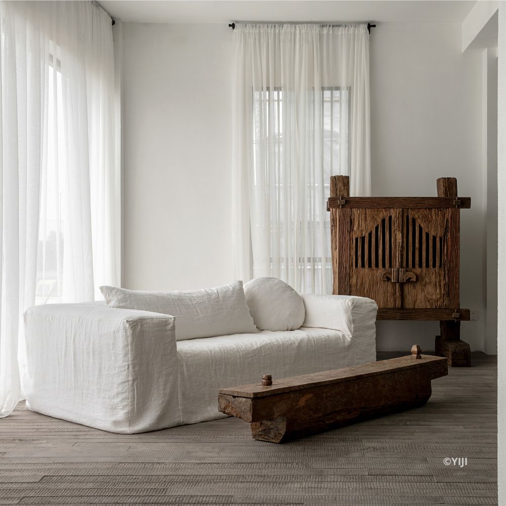 ספה פשתן מבית Fine Lab Home עם כריות לבנות ניצבת בחדר מינימליסטי עם וילונות שקופים, ארון עץ כפרי, ספסל עץ תואם ורצפת עץ בהירה.