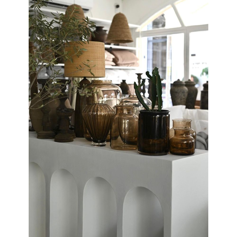 קונסולת קשתות נורדית מבית Fine Lab Home בצבע לבן, עם פרטים מעוגלים, מעוטרת באגרטל זכוכית חום, קקטוס בעציץ כהה, ענף זית ומנורה עם אהיל קלוע בחדר מואר על רצפת בטון מוחלק.