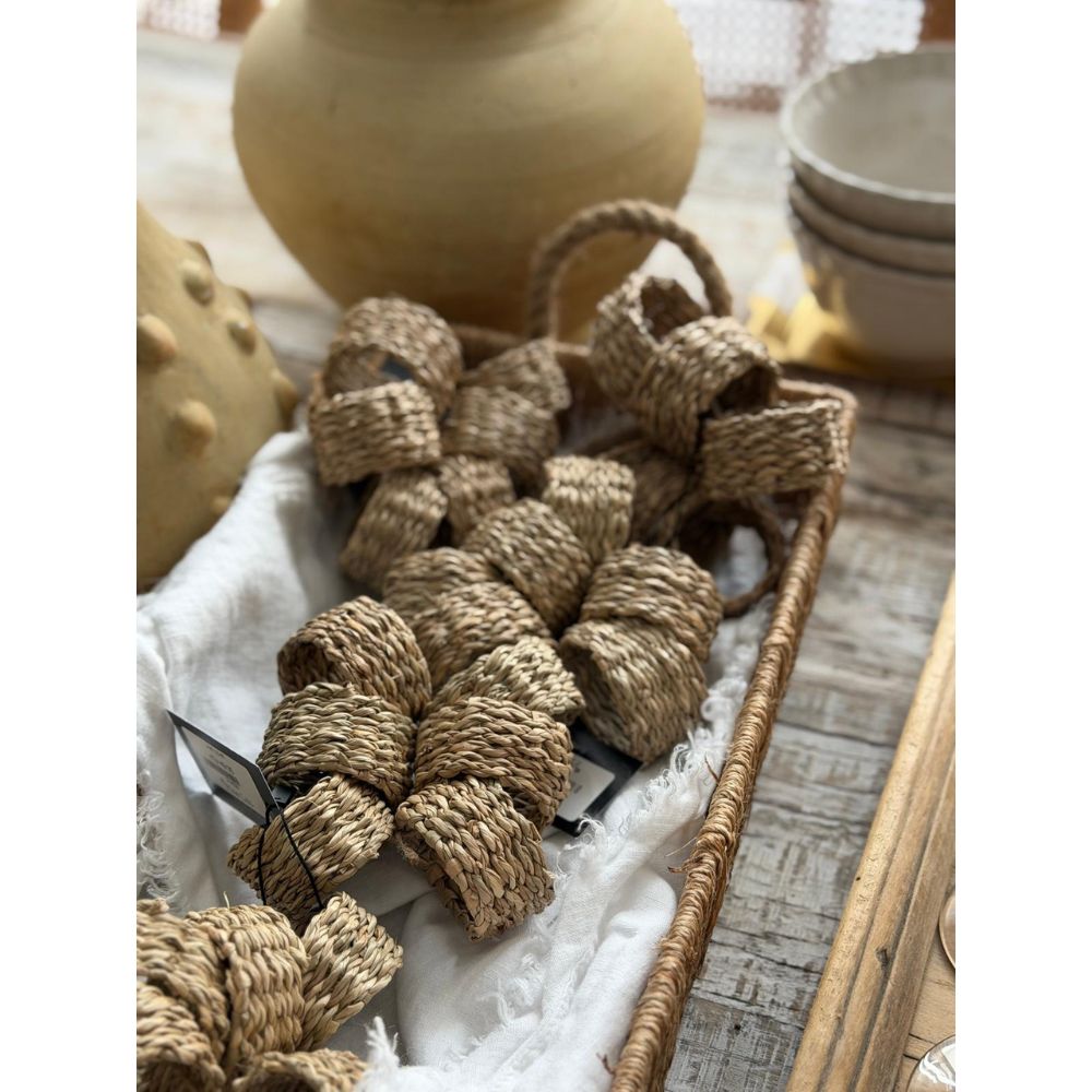 סלסלה קלועה מכילה סט של 6 חובקי מפיות מחבל טבעי מבית Fine Lab Home, המוצגים על מפה לבנה לצד קערות ואגרטלים קרמיים מעשב ים על שולחן עץ.