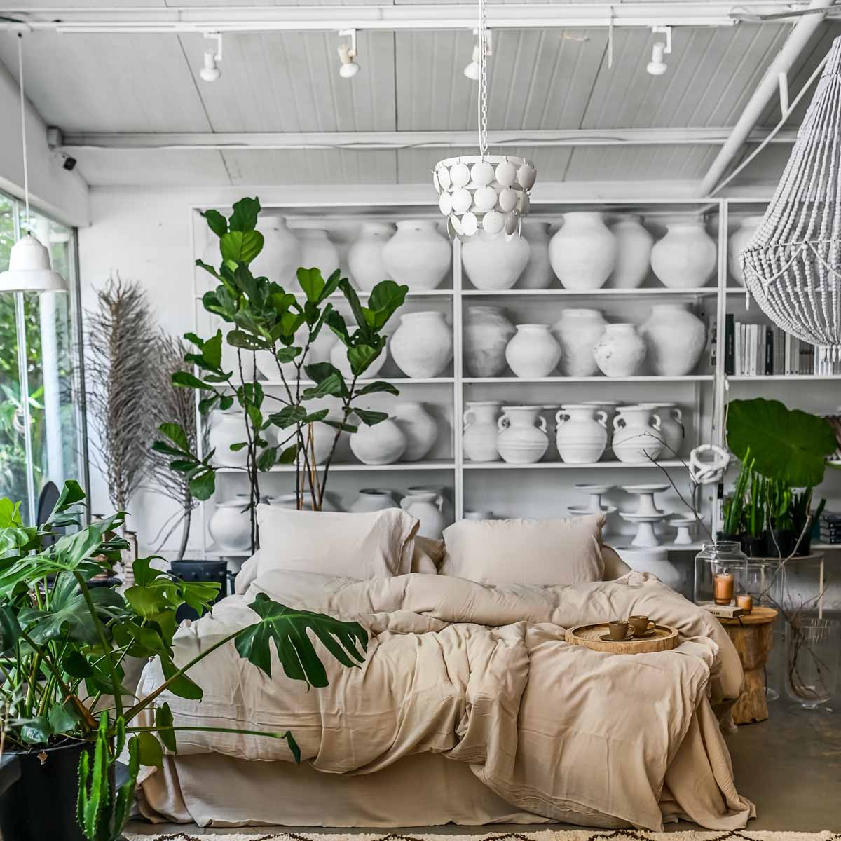 מיטה נעימה עם סט מצעים טטרה חול מבית Fine Lab Home ניצבת מול מדפים עם אגרטלי קרמיקה לבנים, מוקפת צמחים ירוקים בחדר מואר.