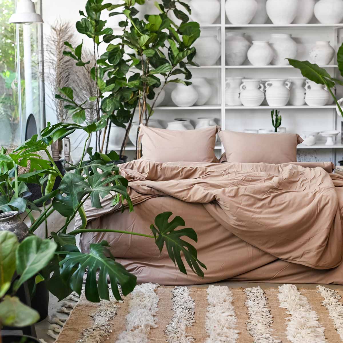 מיטה עטופה בציפה ג'רסי יחיד - שוקולד של Fine Lab Home מוקפת בצמחייה ירוקה ושופעת, ומאחוריה מדפים לבנים עם כלי חרס ואגרטלים. שטיח מרקם בצבעי בז' ולבן משלים את התמונה מול המיטה.