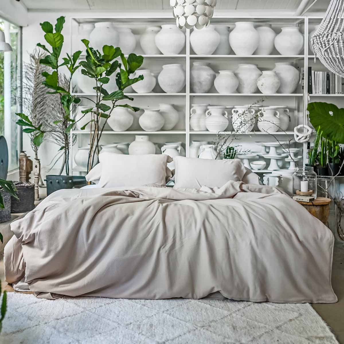 מיטה מסודרת עם מצעי Fine Lab Home's ציפה ג'רסי - אבן מונחת לפני מדפים של כלי חרס לבנים, מוקפת בצמחים ירוקים ושופעים.