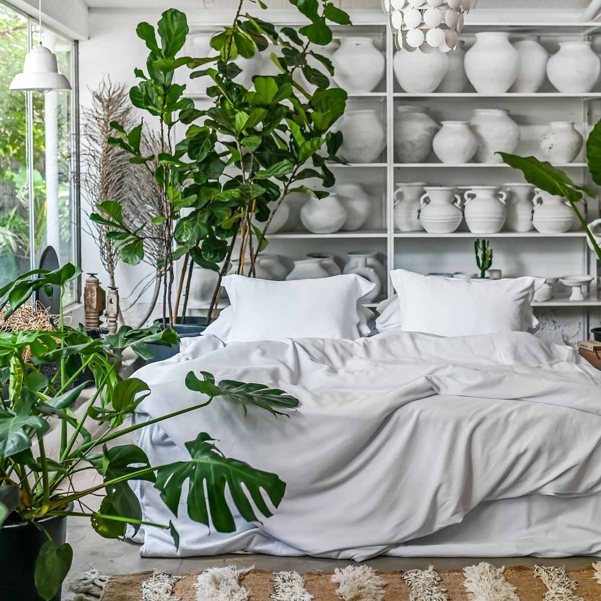 מיטה עם ציפה ג'רסי - לבן מ-Fine Lab Home מכותנה אורגנית מוקף בצמחים ירוקים, לצד מדפים עם אגרטלי קרמיקה לבנים בחדר מודרני ומואר.